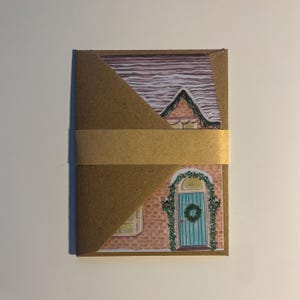 May include: A brown paper card with a watercolor illustration of a house. The house has a teal door with a wreath, a brick facade, and a snow-covered roof. A gold band wraps around the card.