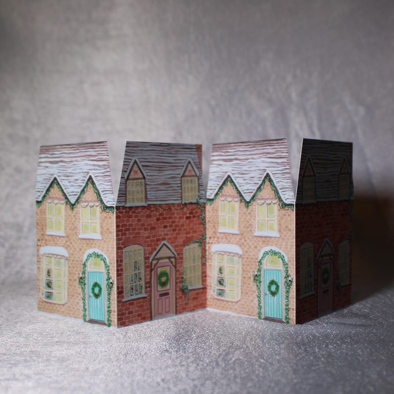 May include: Three paper houses with brick and beige facades, snow-covered roofs, and wreaths on the doors. The windows are lit, suggesting a cozy, festive scene. A charming holiday decoration.
