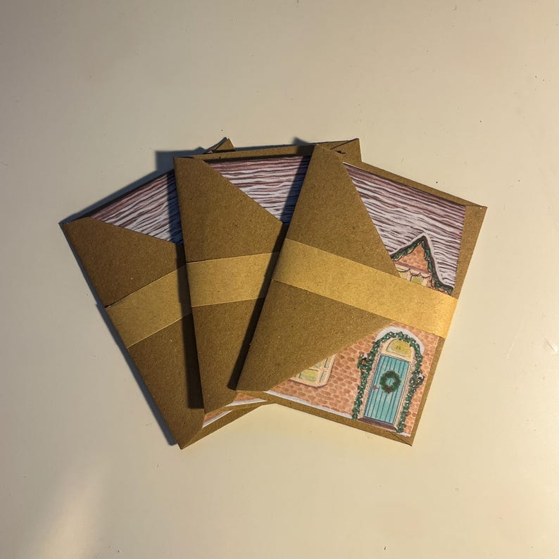 May include: Three folded brown paper cards with a watercolor illustration of a house. Each card is wrapped with a tan paper band. The house illustration features a blue door with a wreath and a pink brick exterior.
