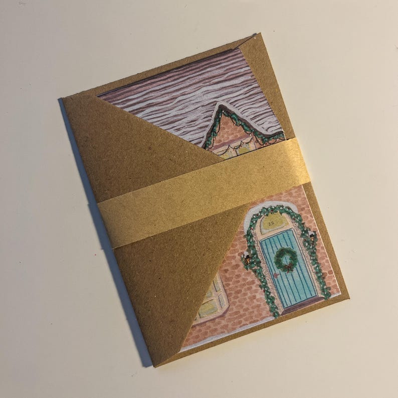 May include: A brown paper envelope with a gold band, holding two illustrated cards. One card shows a house roof with snow, the other a blue door with a wreath. The cards are Christmas themed.