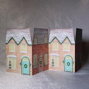 May include: Three-dimensional paper houses with snow-covered roofs and festive wreaths above the doors. The houses are painted in shades of beige and red brick, with light green wreaths and teal doors. The windows have a warm, inviting glow.