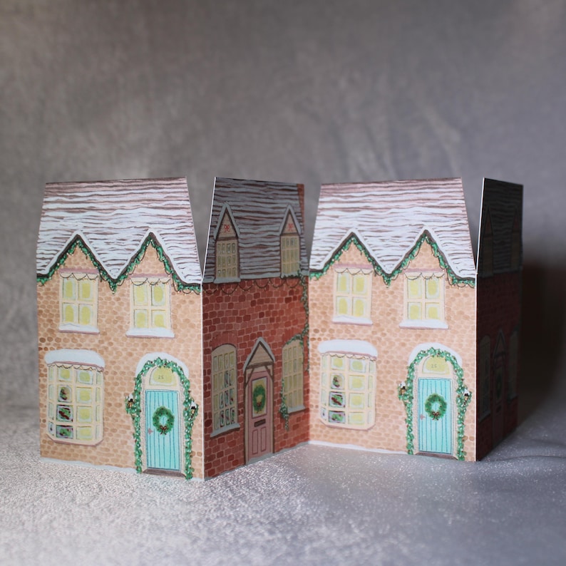 May include: Three-dimensional paper houses with snow-covered roofs and festive wreaths above the doors. The houses are painted in shades of beige and red brick, with light green wreaths and teal doors. The windows have a warm, inviting glow.