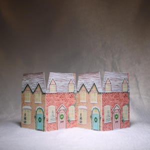 May include: A decorative paper village scene featuring four connected houses. The houses have red brick walls, white-trimmed windows, and gray shingled roofs dusted with snow. Each house has a different colored door and a wreath.