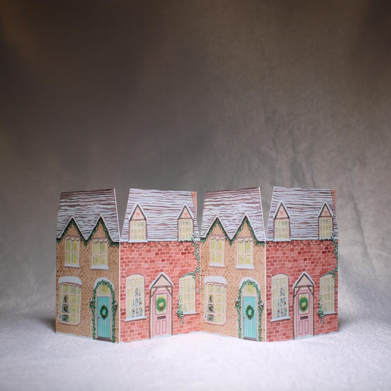 May include: A decorative paper village scene featuring four connected houses. The houses have red brick walls, white-trimmed windows, and gray shingled roofs dusted with snow. Each house has a different colored door and a wreath.
