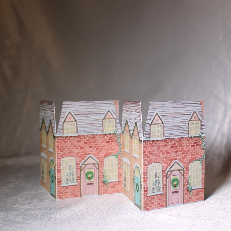 May include: Two paper houses with red brick facades, gray roofs, and small windows. Each house has a different colored door, one pink and one teal, and a wreath on the door. The houses are decorated with string lights and climbing vines.