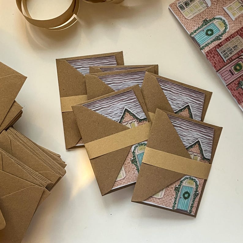 May include: Kraft paper greeting cards with house illustrations, secured with a matching paper band. The cards feature a watercolor-style image of a house with a blue door and a wreath. A stack of blank envelopes is visible.