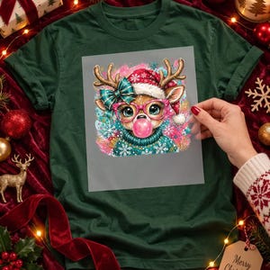 May include: A dark green t-shirt featuring a graphic print of a reindeer wearing a red Santa hat, a teal bow, and pink glasses, while blowing a pink bubble. The reindeer is dressed in a teal sweater with white snowflake patterns.
