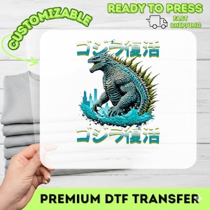 May include: A white DTF transfer featuring a teal Godzilla illustration with Japanese text. The design includes a cityscape and water effects. The transfer is labeled "Customizable" and "Premium DTF Transfer."