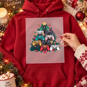 May include: A red hooded sweatshirt featuring a graphic print of a Christmas tree constructed from various colorful video game controllers. The controllers are arranged in a triangular shape, topped with a yellow star, and decorated with small holiday icons.