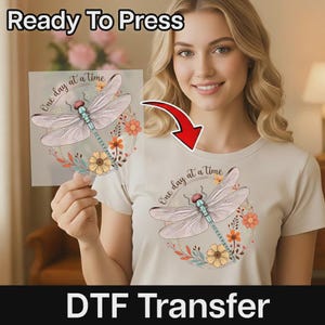 May include: A beige t-shirt featuring a graphic print of a dragonfly with light blue and pink wings, surrounded by orange and yellow flowers and the text One day at a time. A matching transparent transfer sheet is held up next to the shirt.