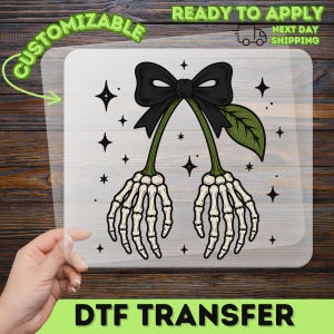 May include: A DTF transfer sheet featuring a graphic of two skeleton hands hanging from a single black bow. The design includes green stems, a green leaf, and several small black four-pointed stars scattered around the central illustration.