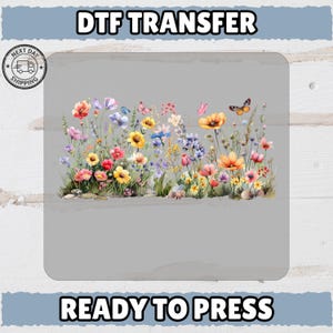 May include: A colorful floral DTF (Direct to Film) transfer design featuring a variety of wildflowers and butterflies. The design includes text that reads "DTF TRANSFER" at the top and "READY TO PRESS" at the bottom. A "NEXT DAY SHIPPING" logo is in the upper left corner.