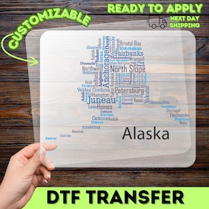 May include: A clear, rectangular DTF transfer featuring a map of Alaska with city names in blue and white. The word "Alaska" is in a large font. The words "Customizable" and "Ready to Apply" are also visible.