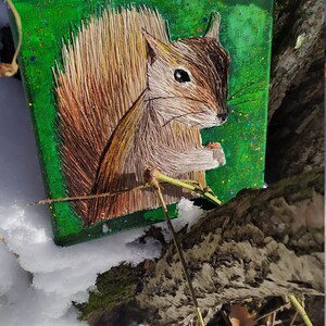 May include: A hand-stitched embroidery of a squirrel on a green canvas with a speckled texture. The squirrel is brown and white with a bushy tail.