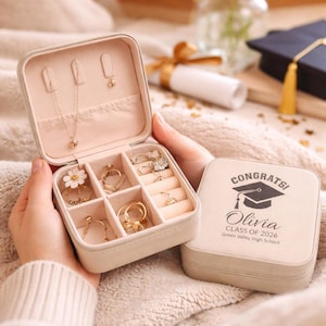 Könnte beinhalten: Eine beige Schmuckschatulle, geöffnet, um goldfarbene Ringe, Halsketten und Ohrringe zu zeigen. Der Innenraum ist rosa. Der geschlossene Deckel ist mit "Congrats!", dem Namen "Olivia" und einer Abschlusskappe bedruckt. Zum Aufbewahren und Schützen von Schmuck.