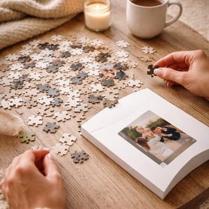 May include: A custom photo puzzle with pieces scattered on a wooden table. A white mug of coffee, a lit candle, and a folded blanket are also on the table. The puzzle's image is visible on the box.