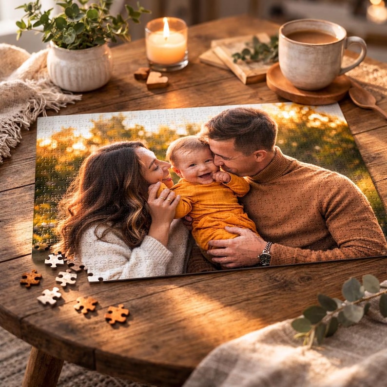 May include: A completed family photo puzzle featuring a smiling baby held by parents. The puzzle is on a wooden table with scattered puzzle pieces. A cup of coffee, a candle, and a plant are also on the table.
