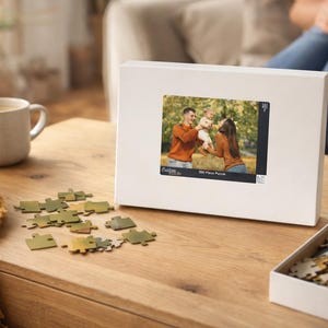 May include: A white puzzle box with a family photo, a partially assembled puzzle, and scattered pieces on a wooden table. The box features a family photo and the text "500 Piece Puzzle". A mug of coffee and a yellow blanket are also visible.