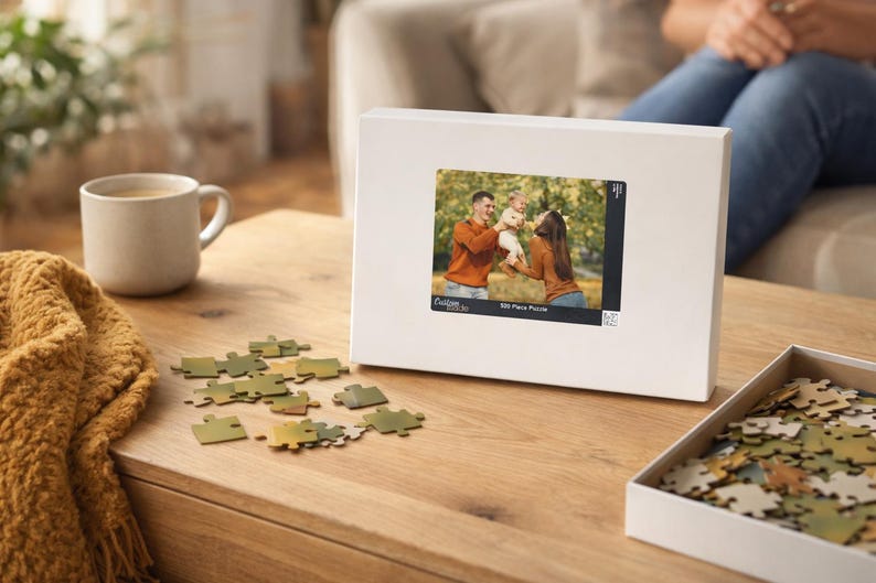 May include: A white puzzle box with a family photo, a partially assembled puzzle, and scattered pieces on a wooden table. The box features a family photo and the text "500 Piece Puzzle". A mug of coffee and a yellow blanket are also visible.