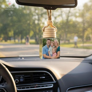 May include: A clear, square car air freshener with a family photo inside, hanging from a rearview mirror. The freshener has a wooden top and a braided rope. The image shows a family of four. The text "Cheerful Artistry" is in the upper left corner.