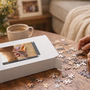 May include: A white puzzle box with an image of a puzzle on the front, next to scattered puzzle pieces on a wooden table. A person is assembling the puzzle. A cup of coffee and a vase of flowers are also on the table.