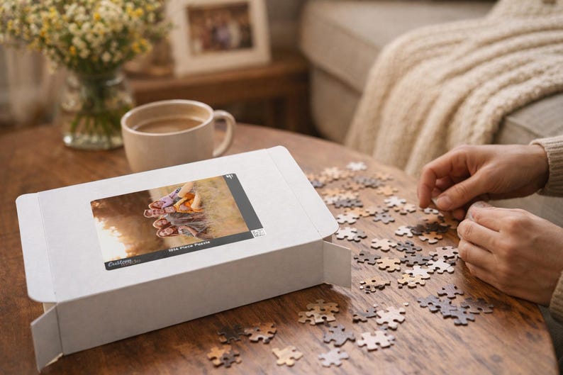 May include: A white puzzle box with an image of a puzzle on the front, next to scattered puzzle pieces on a wooden table. A person is assembling the puzzle. A cup of coffee and a vase of flowers are also on the table.