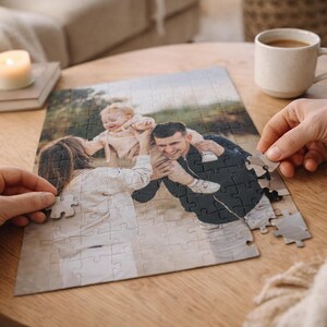 May include: A family photo puzzle in progress, featuring a smiling couple and a baby. The puzzle is on a wooden table, with hands placing the pieces. A lit candle and a coffee cup are also on the table, creating a cozy atmosphere.