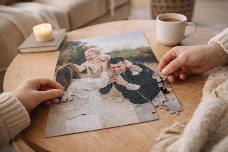 May include: A family photo puzzle in progress, featuring a smiling couple and a baby. The puzzle is on a wooden table, with hands placing the pieces. A lit candle and a coffee cup are also on the table, creating a cozy atmosphere.