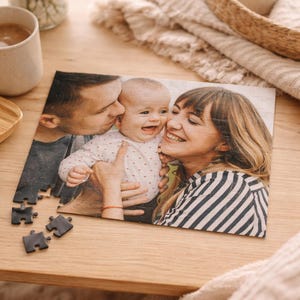May include: A personalized photo puzzle featuring a family portrait. The image shows a smiling baby being kissed by two adults. Several puzzle pieces are separate from the assembled image, resting on a wooden surface.