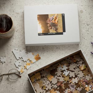 May include: A still-life image featuring a white puzzle box with a photo of people, a partially assembled puzzle, a box of puzzle pieces, a cup of tea, glasses, and lavender sprigs. The puzzle box has the text "255 Piece Puzzle".