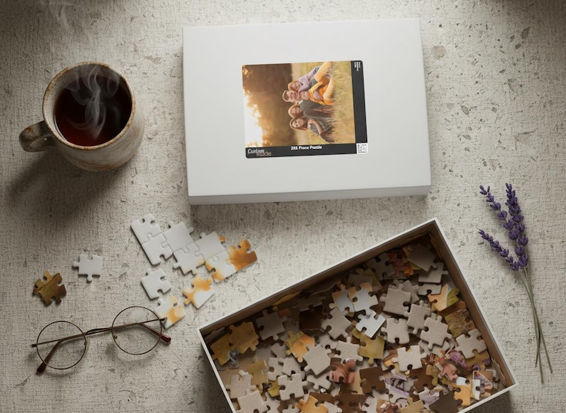 May include: A still-life image featuring a white puzzle box with a photo of people, a partially assembled puzzle, a box of puzzle pieces, a cup of tea, glasses, and lavender sprigs. The puzzle box has the text "255 Piece Puzzle".