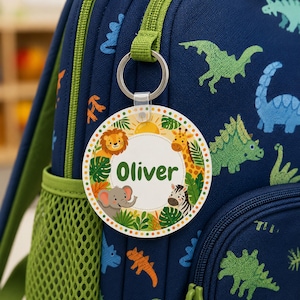 May include: A circular white plastic bag tag featuring the name Oliver in green text, surrounded by illustrations of a lion, giraffe, elephant, and zebra amidst tropical leaves, attached to a navy blue backpack with a dinosaur pattern.