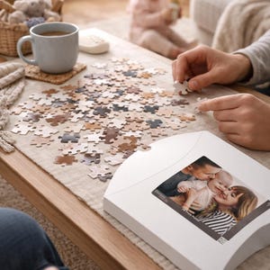 May include: A family is working on a puzzle on a wooden coffee table. The puzzle pieces are various shades of brown and beige. A white puzzle box with a family photo is on the table. A cup of tea and a baby are also in the scene.