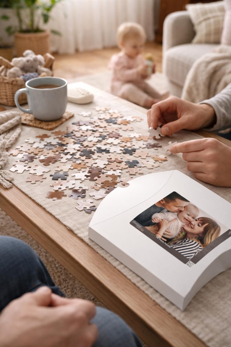 May include: A family is working on a puzzle on a wooden coffee table. The puzzle pieces are various shades of brown and beige. A white puzzle box with a family photo is on the table. A cup of tea and a baby are also in the scene.