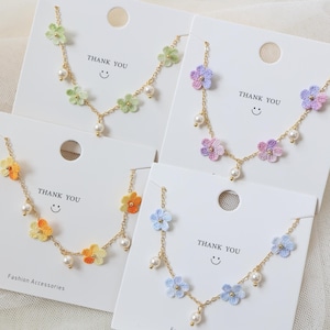 Könnte beinhalten: Eine Auswahl an Blumenketten in verschiedenen Farben, darunter Grün, Gelb, Blau und Lila. Jede Halskette hat kleine, gestickte Blumen und baumelnde Kunstperlen an einer goldfarbenen Kette. Die Halsketten werden auf weißen Karten mit dem Aufdruck "THANK YOU" präsentiert.