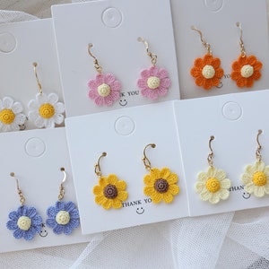 May include: Six pairs of crocheted flower earrings displayed on white cards. The earrings are in various colors, including white, yellow, orange, pink, and blue. Each pair features a crocheted flower with a small yellow center.
