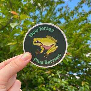 Pode incluir: Um autocolante redondo com uma ilustra&ccedil;&atilde;o de um sapo amarelo e rosa. O texto "New Jersey" e "Pine Barrens" est&aacute; disposto &agrave; volta do sapo sobre um fundo verde escuro. O autocolante &eacute; mostrado sobre um fundo de folhagem verde.