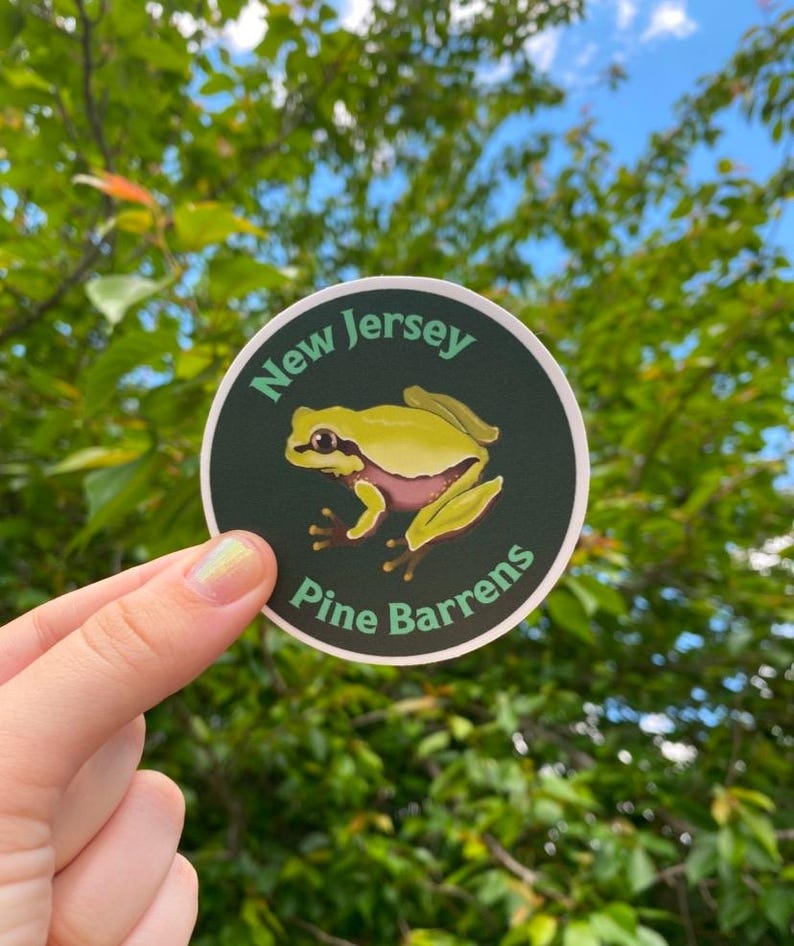 Pode incluir: Um autocolante redondo com uma ilustra&ccedil;&atilde;o de um sapo amarelo e rosa. O texto "New Jersey" e "Pine Barrens" est&aacute; disposto &agrave; volta do sapo sobre um fundo verde escuro. O autocolante &eacute; mostrado sobre um fundo de folhagem verde.