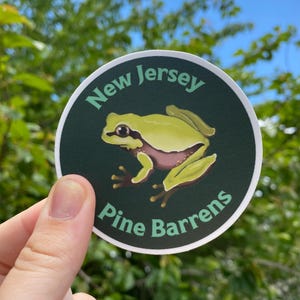 Pode incluir: Um autocolante redondo com uma ilustra&ccedil;&atilde;o de um sapo-verde e o texto "New Jersey Pine Barrens". O texto e o sapo est&atilde;o sobre um fundo verde escuro com uma borda branca.