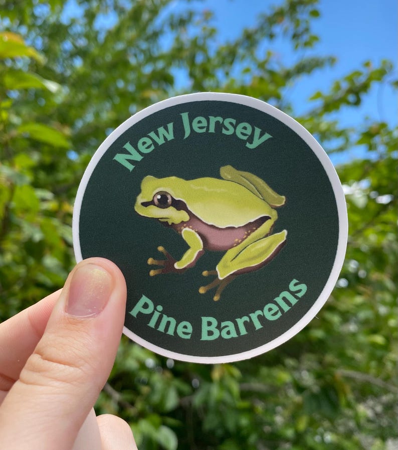 Pode incluir: Um autocolante redondo com uma ilustra&ccedil;&atilde;o de um sapo-verde e o texto "New Jersey Pine Barrens". O texto e o sapo est&atilde;o sobre um fundo verde escuro com uma borda branca.