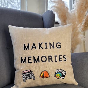 May include: A square beige throw pillow with a textured fabric finish. The text "MAKING MEMORIES" is printed in black capital letters. Below the text are three small illustrations: a black jeep, an orange camping tent, and a black camera.