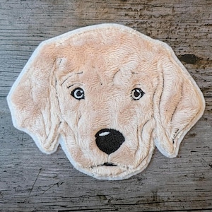 Könnte beinhalten: Beiger Hundekopf-Teppich mit schwarzer Nase und detaillierten Augen. Der Teppich hat ein weiches, strukturiertes Aussehen. Der Hintergrund ist eine Holzoberfläche mit sichtbarer Maserung.