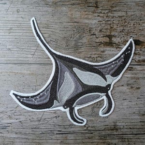Könnte beinhalten: Gestickter Aufnäher mit einem Mantarochen-Design. Der Aufnäher zeigt Grau- und Schwarztöne mit weißen Akzenten und einem weißen Rand. Der Aufnäher befindet sich auf einer verwitterten Holzoberfläche.