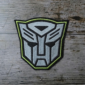 Può includere: Toppa ricamata con il simbolo Autobot. La toppa ha un design grigio e nero con un bordo verde neon. La toppa è su una superficie di legno invecchiato.