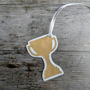 May include: A gold-colored trophy-shaped ornament with a white border and a white ribbon for hanging. The ornament is made of felt and is set against a weathered wood background.
