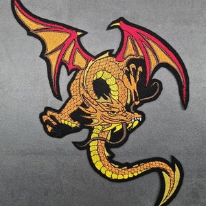 Könnte beinhalten: Ein gestickter Bügel-Patch mit einem gelben und orangefarbenen Drachen mit roten Flügeln. Der Drache ist in einer zusammengerollten Position mit ausgebreiteten Flügeln.