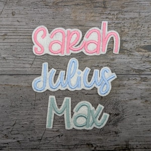 May include: Three embroidered name patches: "Sarah" in pink, "Julius" in light blue, and "Max" in teal. Each name is stitched onto a white felt background. The patches are displayed on a weathered, gray wooden surface.