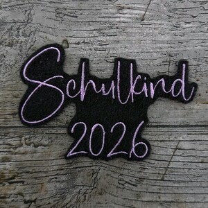 以下が含まれることがあります： ラベンダー色で刺繍された「Schulkind 2026」の文字が入った黒いパッチ。 パッチはユニークな形をしており、灰色の木製の背景に配置されています。