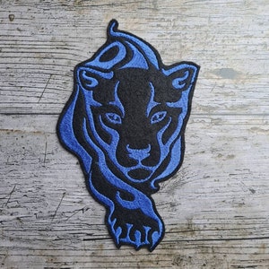 May include: Black panther head embroidered patch with blue outline. The patch is on a light wood background.