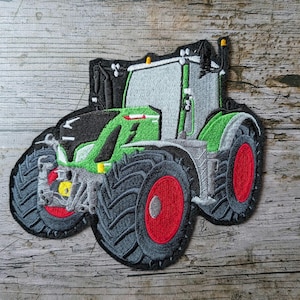 May include: Embroidered patch of a green, gray, and black tractor with red wheels. The tractor has detailed stitching and is set against a wooden background. The patch is designed to be ironed or sewn onto fabric.